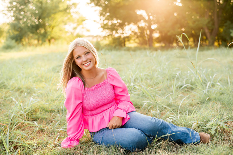 High school senior girl sits on her hip and smiles in a pink shirt and blue jeans in a field in central Oklahoma for her senior photos