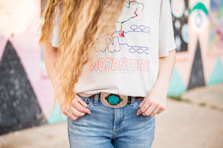 cropped photo of  torso area  of a high school senior girl with long blond hair and hands in her belt loops wearing a big belt buckle and vintage t-shirt and blue jeans standing in front of a mural at Factory Obscura in midtown Oklahoma City