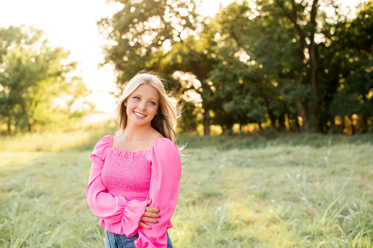 High school senior stands holding one arm in a green field in central Oklahoma for her senior photos