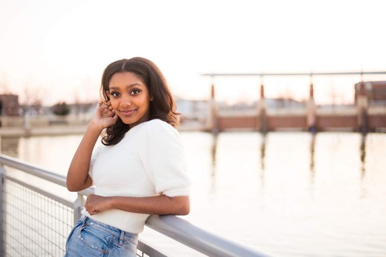 African-American teenage girl leans back against a railing at the waters edge at her cap and gown senior photo shoot in Norman Oklahoma