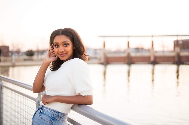 African-American teenage girl leans back against a railing at the waters edge at her cap and gown senior photo shoot in Norman Oklahoma