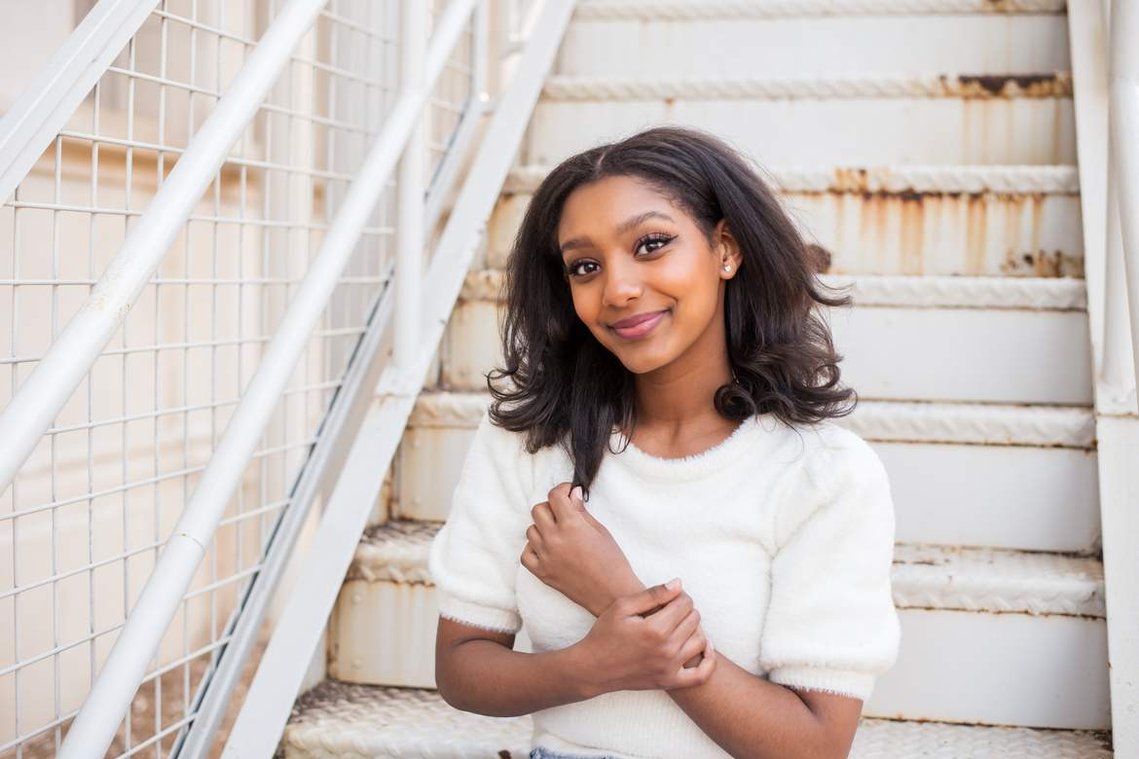 African-American teenage girl sits on white steps in a white sweater and smiles at her cap and gown senior photo shoot in Norman Oklahoma
