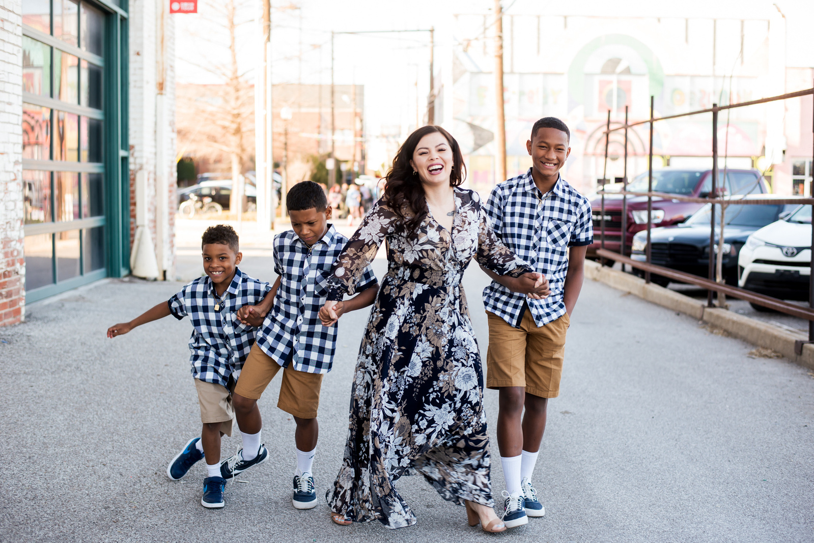 a mom with her 3 young boys are holding hands and walking and smiling in an alley with buildings around them in midtown Oklahoma City
