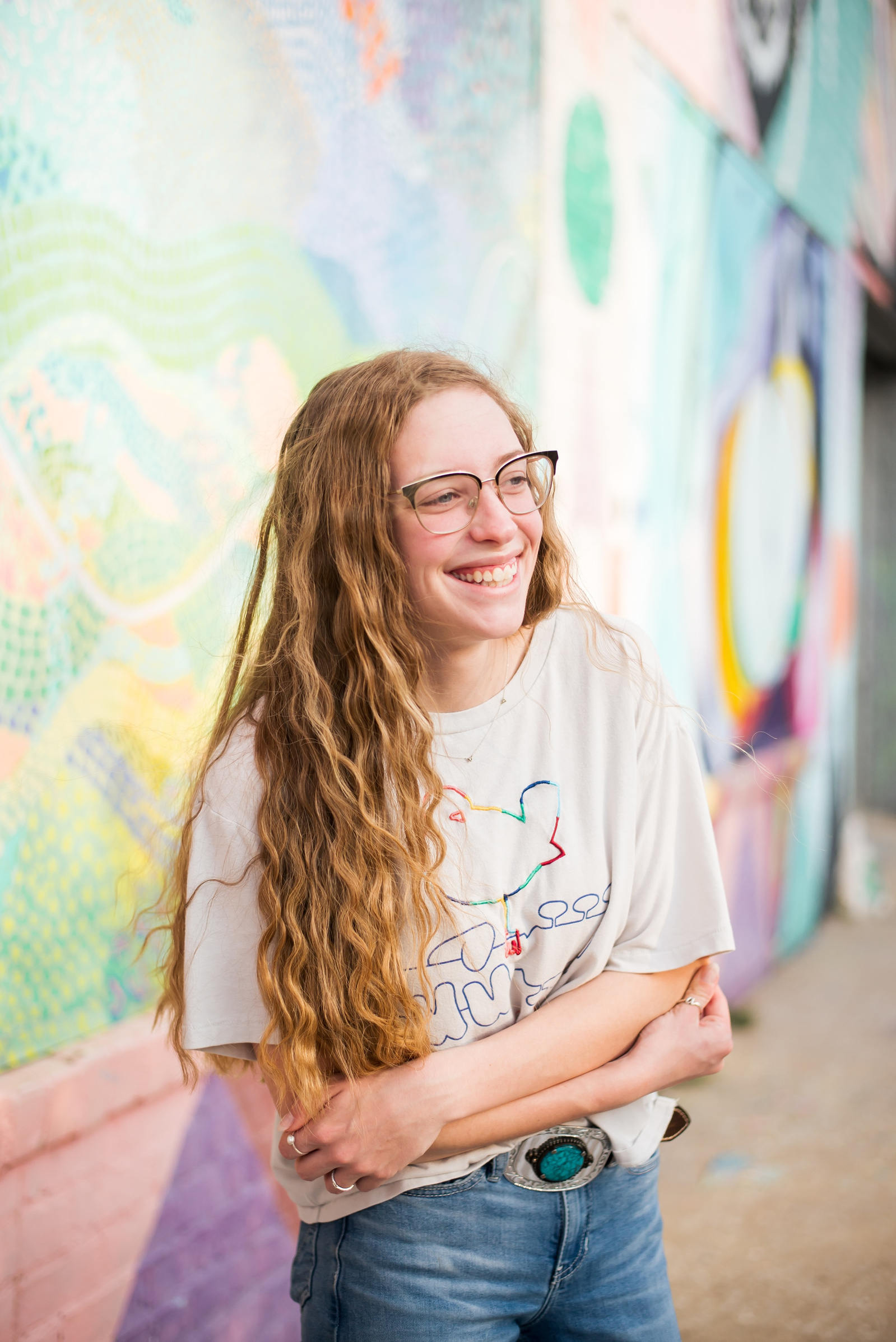 high school senior girl with long blond hair and glasses dressed retro, stands with her arms crossed and smiles off camera in front of murals in Automobile Alley in Oklahoma City