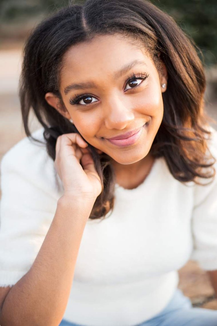 Close up portrait of African-American teenage girl tucking hair behind her ear and smiling in a white sweater at her cap and gown senior photo shoot in Norman Oklahoma