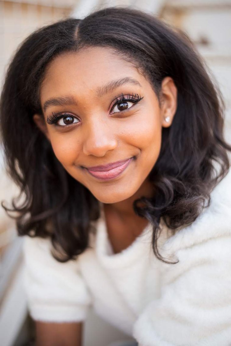 close-up portrait African-American teenage girl sits on white steps in a white sweater and smiles at her cap and gown senior photo shoot in Norman Oklahoma