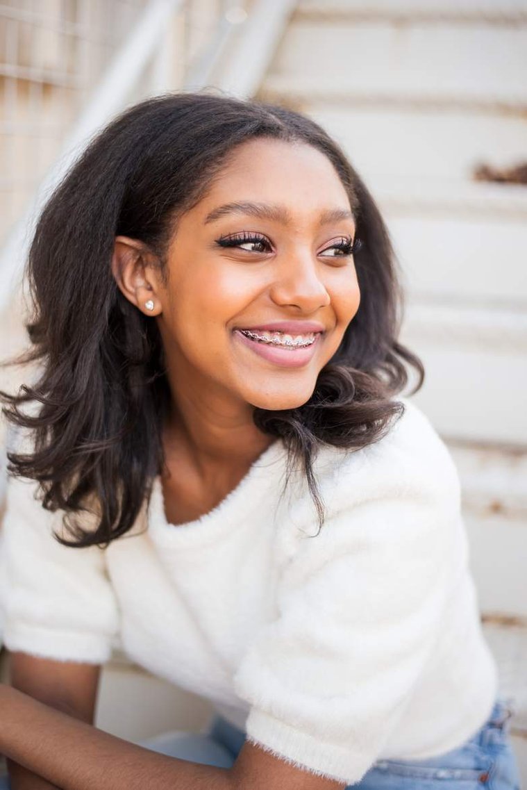 African-American teenage girl sits on white steps in a white sweater and smiles at her cap and gown senior photo shoot in Norman Oklahoma