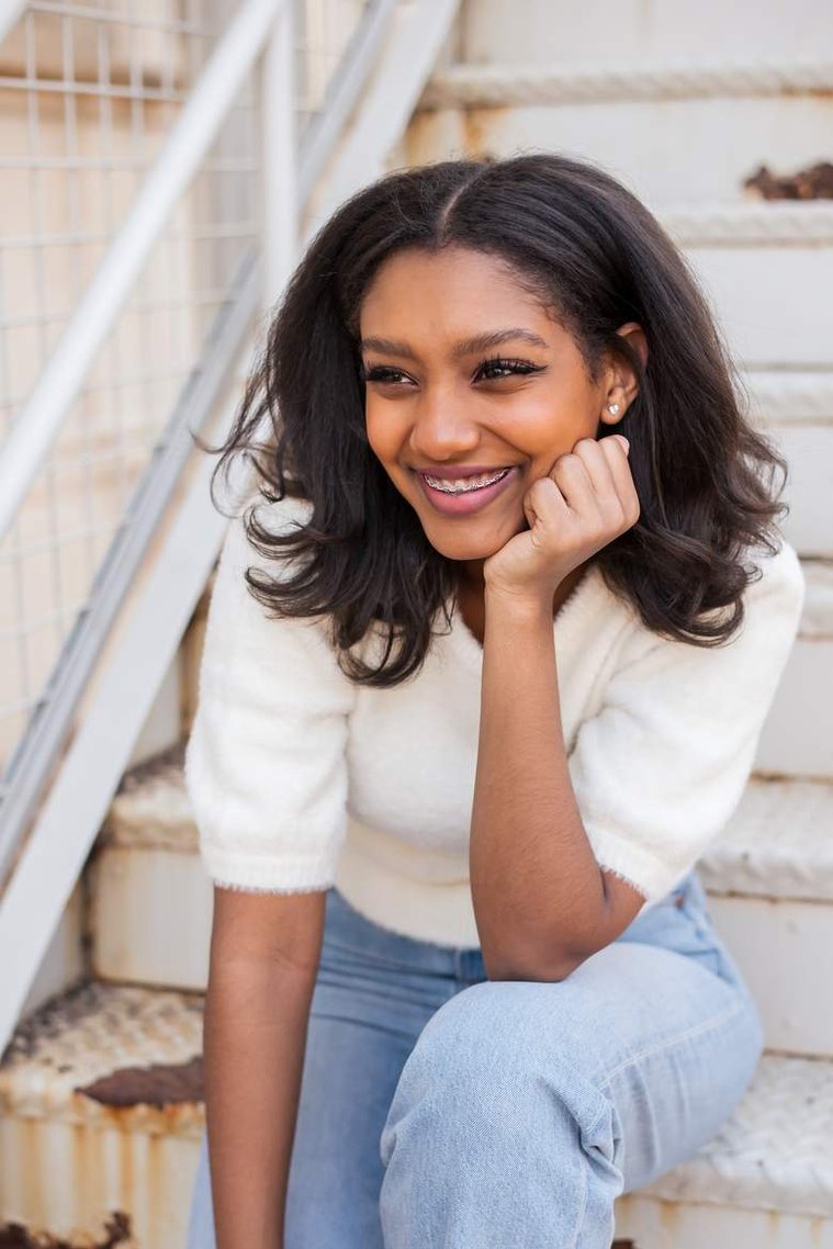 African-American teenage girl sits on white steps in a white sweater and laughs at her cap and gown senior photo shoot in Norman Oklahoma