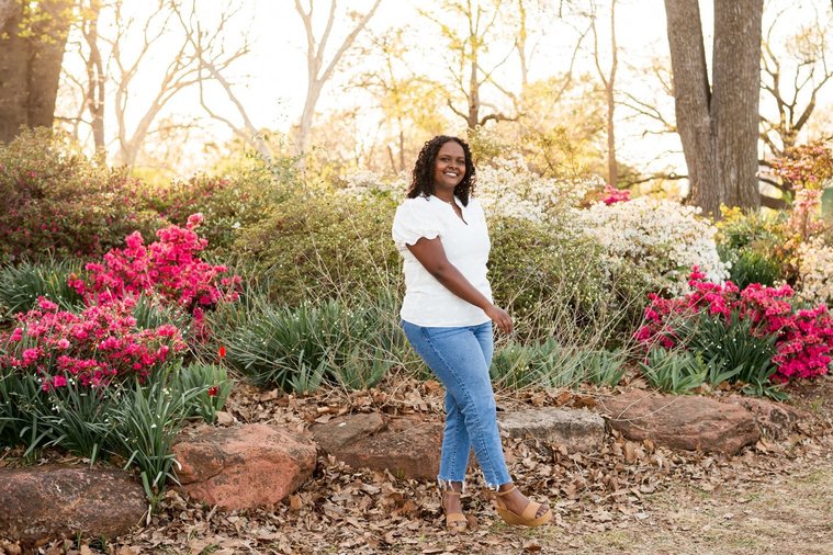 senior girl wearing jeans and a white top stands playfully smiling among the flowers and trees of Will Rogers Gardens in OKC