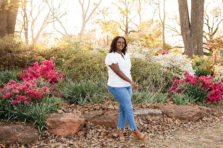 senior girl wearing jeans and a white top stands playfully smiling among the flowers and trees of Will Rogers Gardens in OKC