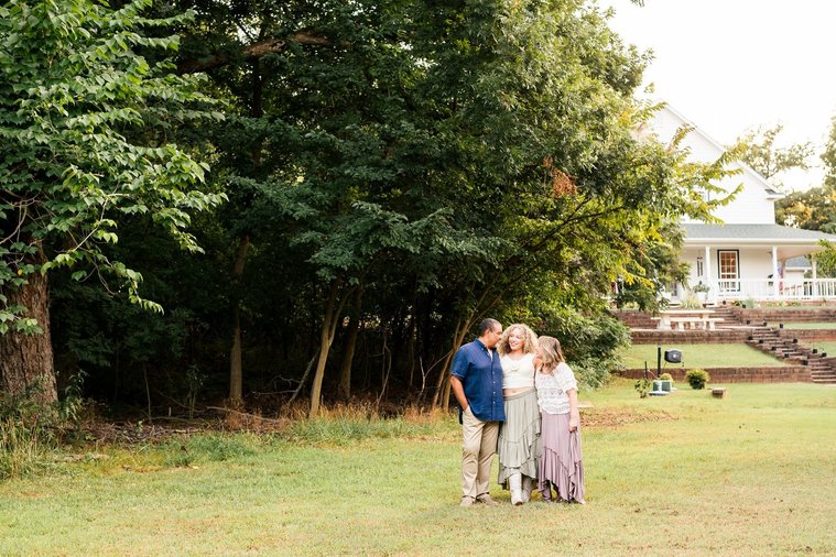 wide angle photo of a mom, dad, and teen daughter standing together talking with big trees and their home behind them in Norman, Oklahoma