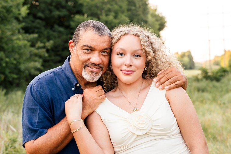 a dad and teen daughter sit together hugging and smiling in a field with trees behind them in Norman, Oklahoma