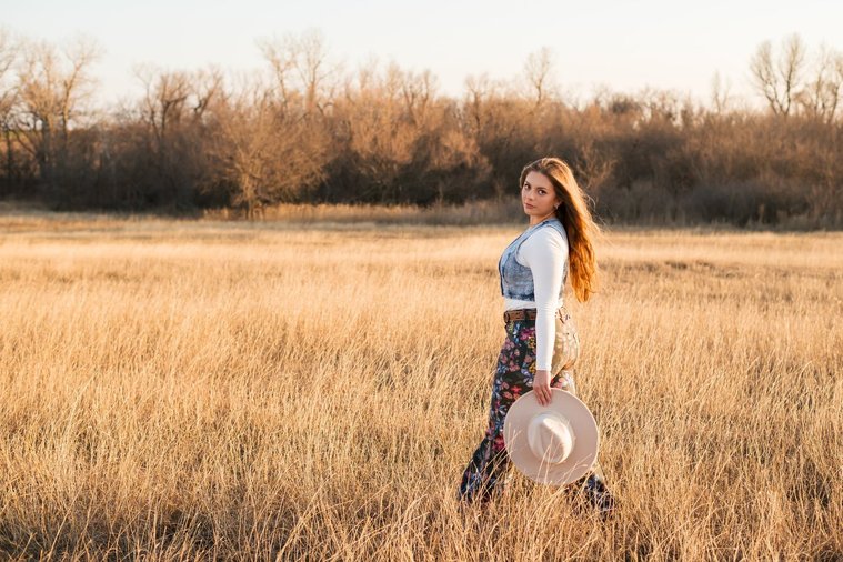 high school senior girl with long brown hair wearing cowboy boots, Wranglers, and a denim vest looks over her shoulder holding her western hat in her hand as she walks through a field of golden grass in the country near OKC