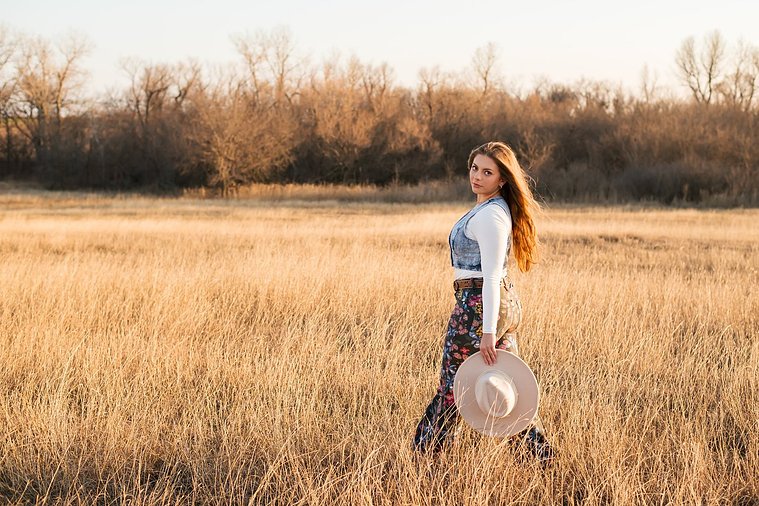 high school senior girl with long brown hair wearing cowboy boots, Wranglers, and a denim vest looks over her shoulder holding her western hat in her hand as she walks through a field of golden grass in the country near OKC