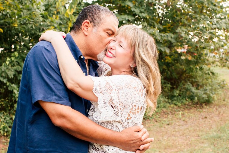 a middle aged husband and wife hug each other smiling and the husband is kissing his wife on the cheek with trees behind them at their home in Norman, Oklahoma