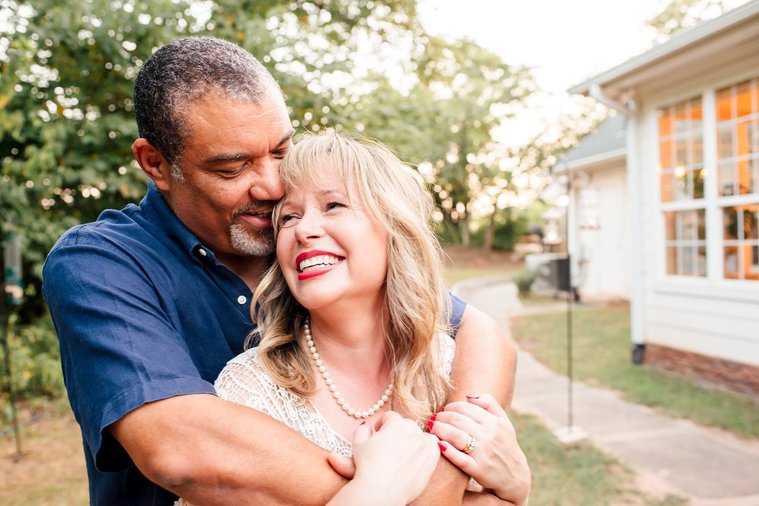 a middle aged husband hugs his wife from behind while she looks up at him lovingly with trees and their home behind them in Norman, Oklahoma