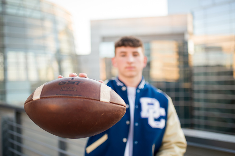 senior boy wearing letterman jacket holds a football close to camera with downtown OKC buildings behind him
