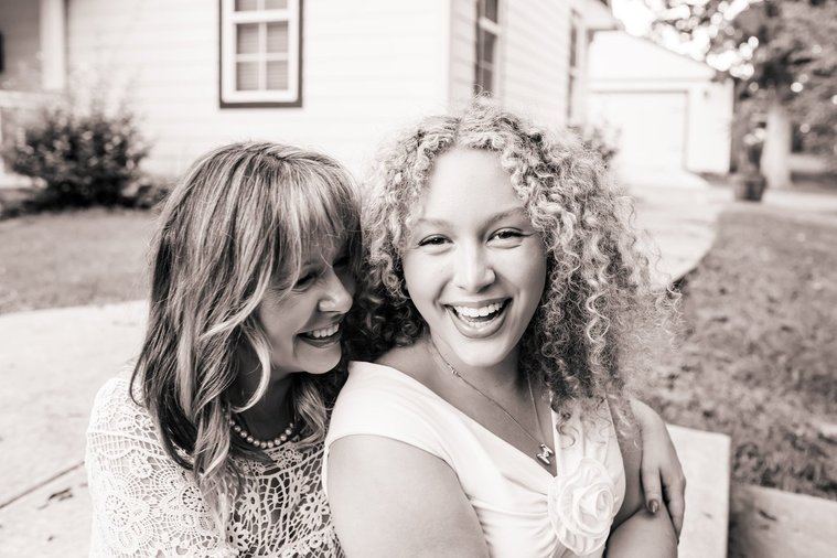 black and white photo of a mom and her teen daughter sitting together on their front sidewalk and laughing intimately with their home behind them in Norman, Oklahoma