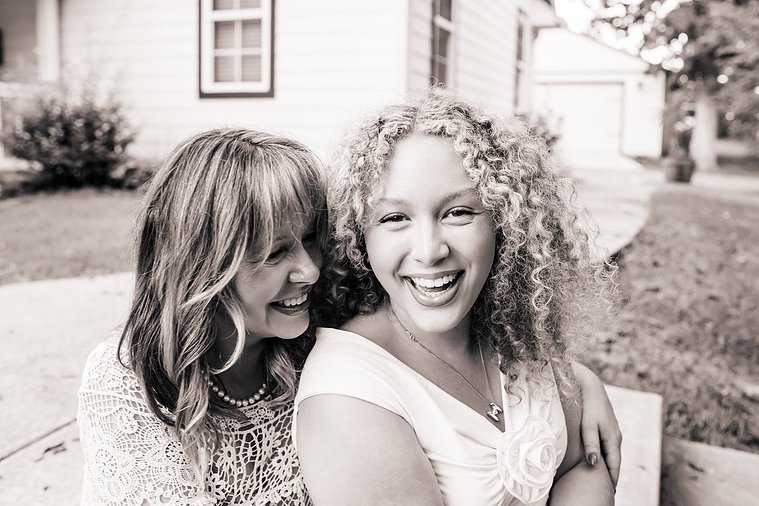 black and white photo of a mom and her teen daughter sitting together on their front sidewalk and laughing intimately with their home behind them in Norman, Oklahoma