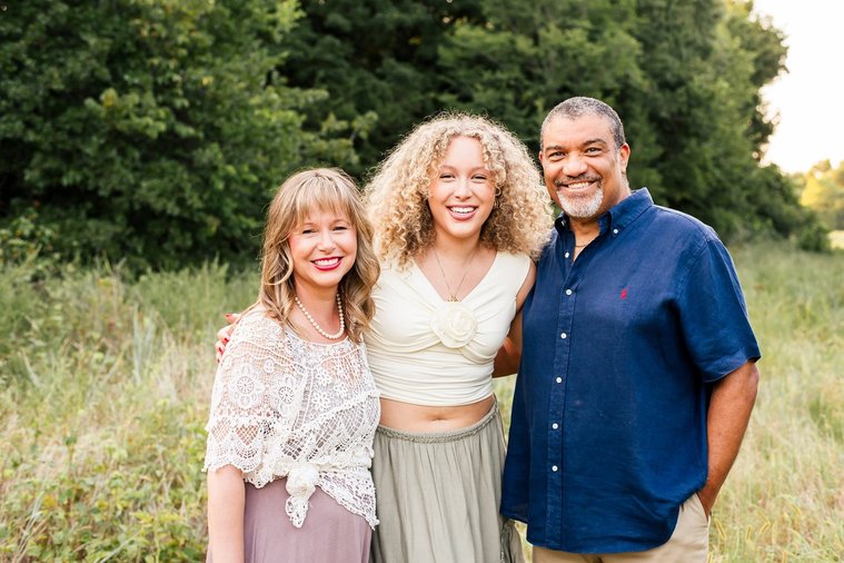 a mom, dad, and teen daughter stand together hugging and smiling in a field with trees behind them in Norman, Oklahoma