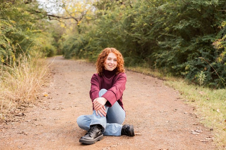 senior girl with red hair wearing a purple sweater and jeans sits cross legged and smiling on a path with fall trees behind her at Sutton Park in Norman, Oklahoma