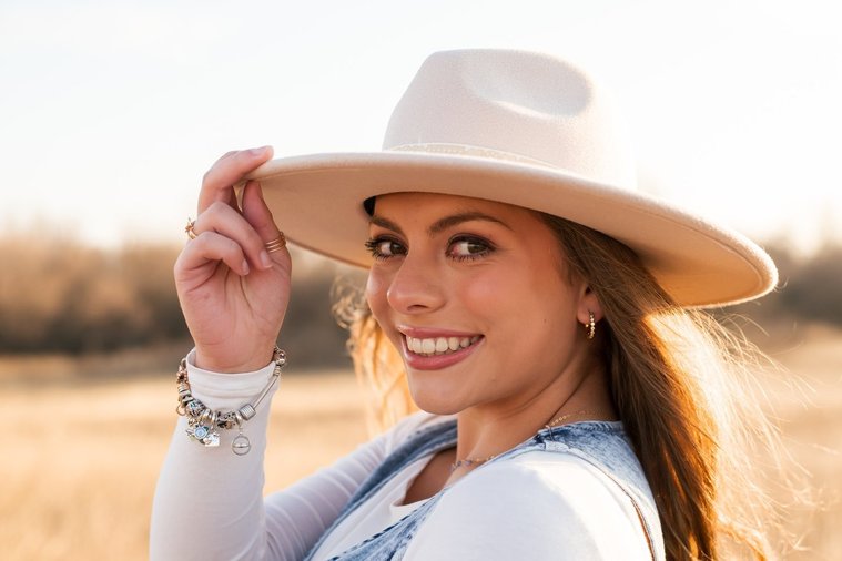 high school senior girl with long brown hair wearing a denim vest and western hat smiles over her shoulder while she holds the brim of her hat standing in a field of golden grass in the country near OKC