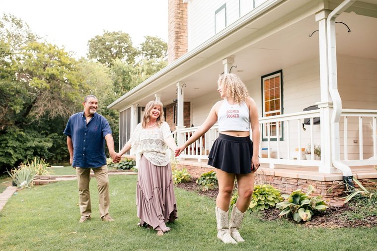 a mom, dad, and teen daughter hold hands in a line with the daughter in front wearing OU college gear and looking back to her parents to represent leaving for college with their porch behind them in Norman, Oklahoma