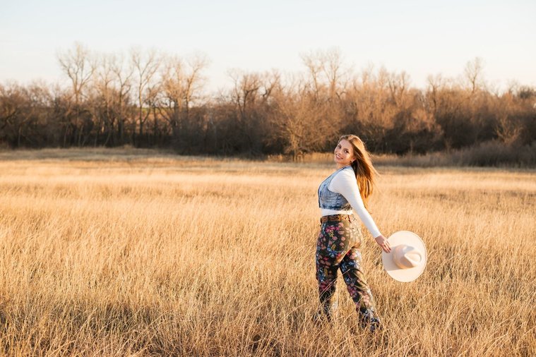 high school senior girl with long brown hair wearing cowboy boots, Wranglers, and a denim vest smiles playfully over her shoulder as she walks through a field of golden grass in the country near OKC holding her western hat behind her