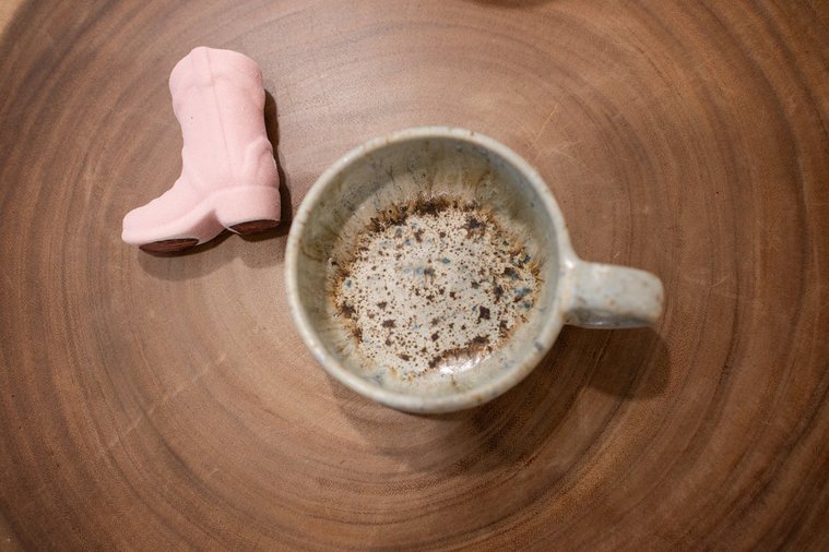 close up branding image of an empty coffee cup with a small pink cowboy boot decor piece next to it against a woodgrain table at a graphic design team branding session in OKC