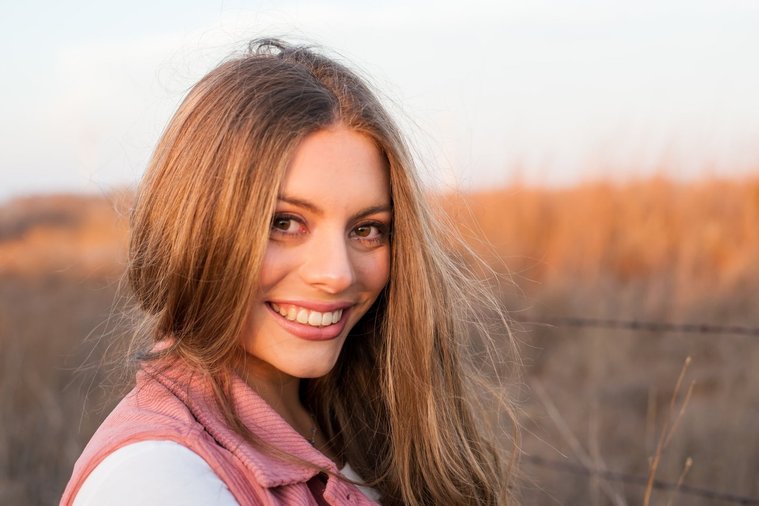 portrait of a beautiful senior girl with long hair wearing a pink vest smiling over her shoulder, standing in front of a country field near Oklahoma City