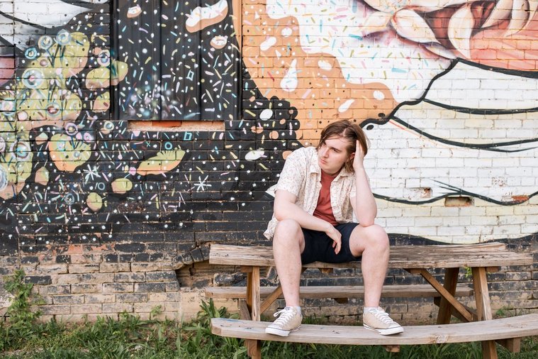senior boy wearing a button down shirt and blue shorts sits on a picnic table with one hand in hair looking away in front of a colorful wall mural in midtown Oklahoma City