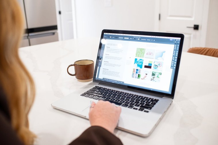 OKC branding photo session image taken over the shoulder of a woman working on graphic design projects sitting at a table with a cup of coffee beside her
