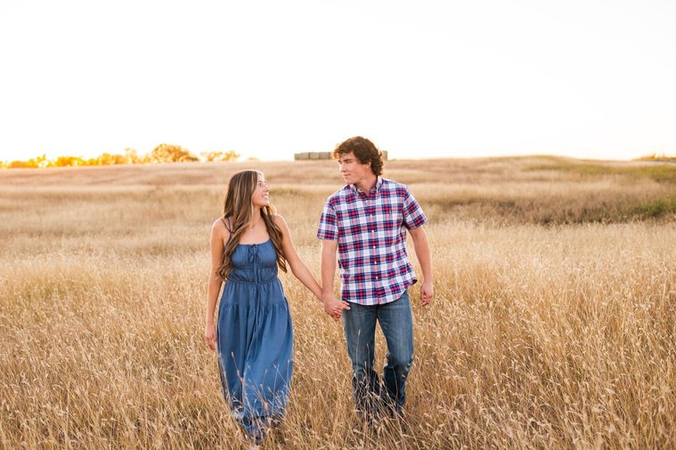 senior girl in a blue dress with her boyfriend walk together in a field in the country outside of Oklahoma City holding hands 