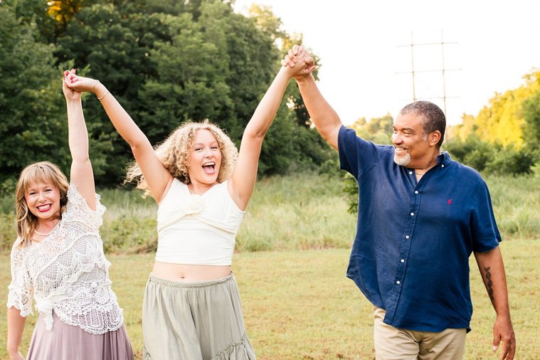 a mom, dad, and teen daughter are walking holding hands while the daughter holds everyone's hands in the air and laughs, in a field with trees behind them in Norman, Oklahoma
