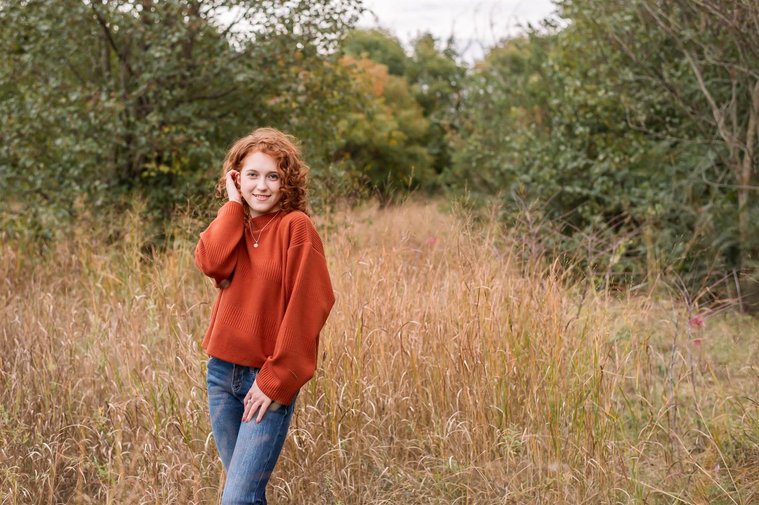 senior girl with red hair wearing an orange sweater and jeans poses standing in a field with gold grass and fall trees behind her at Sutton Park in Norman, Oklahoma
