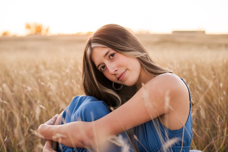 senior girl with long brown hair wearing a blue sundress sits with her knees tucked under her chin smiling softly in a country field in the fall near Oklahoma City