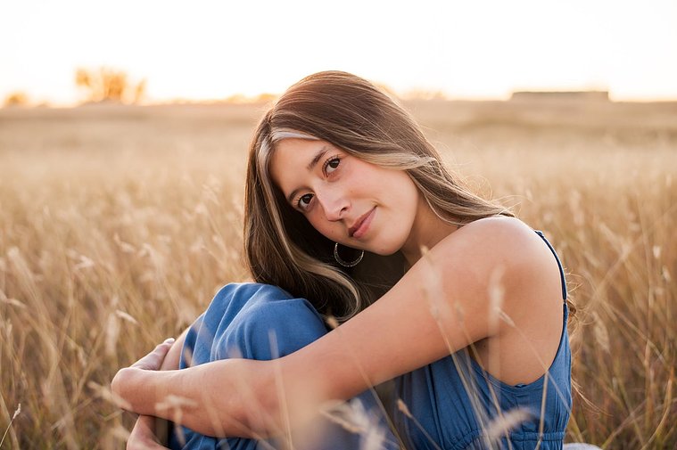 senior girl with long brown hair wearing a blue sundress sits with her knees tucked under her chin smiling softly in a country field in the fall near Oklahoma City