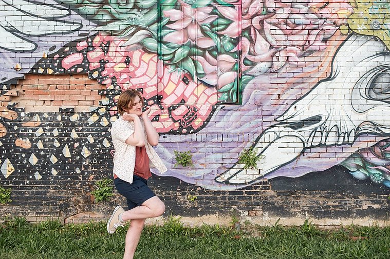 senior boy in a button down shirt and shorts making a funny dramatic face with his leg kicked back in front of a colorful wall mural in midtown Oklahoma City