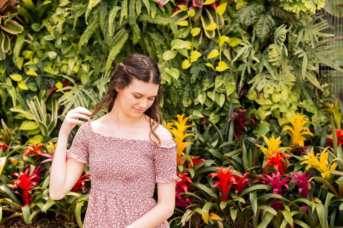 high school senior girl with long brown hair in a red and white floral dress sits in front of a tropical plant wall and looks down softly holding the end of her hair at Crystal Bridge Conservatory at Myriad Gardens in Oklahoma City