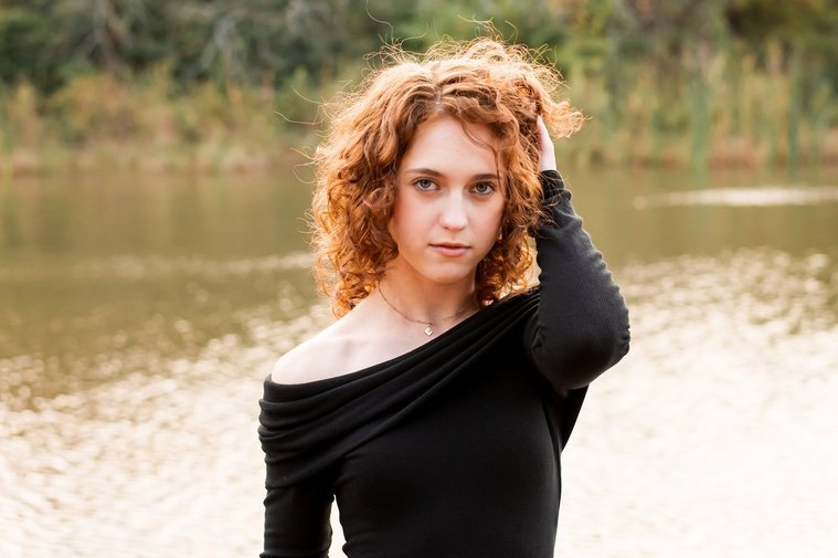senior girl with red hair wearing an elegant long black dress stands near a pond with one hand in her hair and looking at camera with no smile at Sutton Park in Norman, Oklahoma