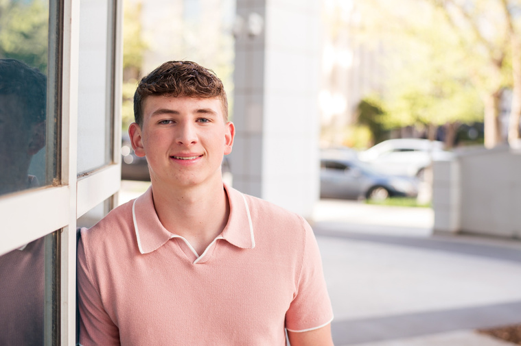 high school senior boy wearing a peach polo top leans against reflective glass windows of the downtown library in Oklahoma City