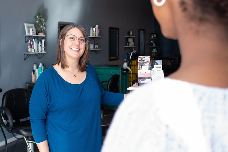 looking over clients shoulder at a woman in a blue shirt and glasses smiles welcomingly at the client entering a salon at a branding session at Vibe Beauty Bar in Tuttle, Oklahoma