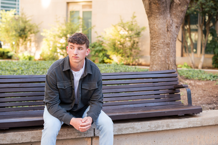 high school senior boy wearing a gray jacket and jeans sits casually on a bench looking to the side in front of a building in downtown OKC
