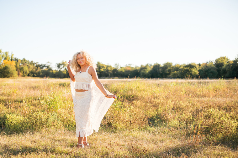 wide angle portrait of a high school senior girl with blonde curly hair wearing a boho western white fringe dress holding a crochet blanket behind her in a field at Martin Nature Park in Oklahoma City