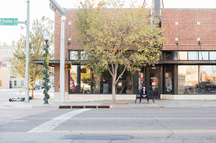 wide angle image of a hispanic high school senior boy wearing a dark blue suit and white shirt sitting on a black bench on the sidewalk with the street sign and road around him in Automobile Alley in Oklahoma City