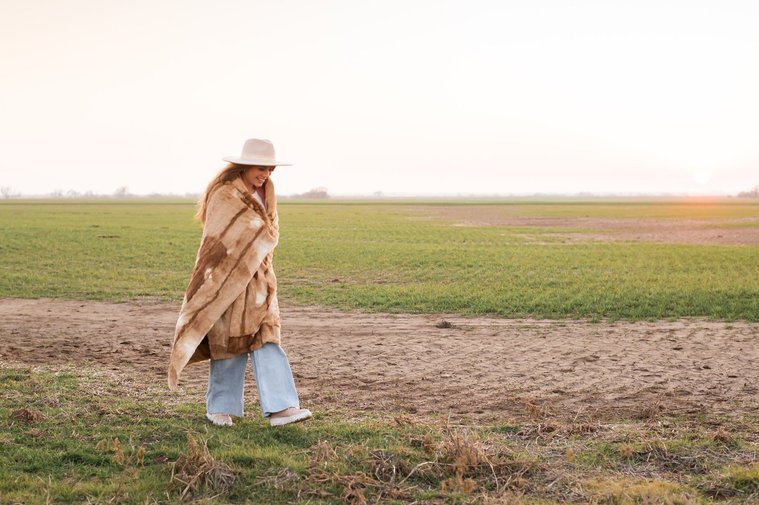 high school senior girl with long brown hair wearing a western hat wrapped in a brown blanket walks across frame looking down in a field near OKC with the sunset behind her