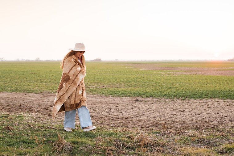 high school senior girl with long brown hair wearing a western hat wrapped in a brown blanket walks across frame looking down in a field near OKC with the sunset behind her