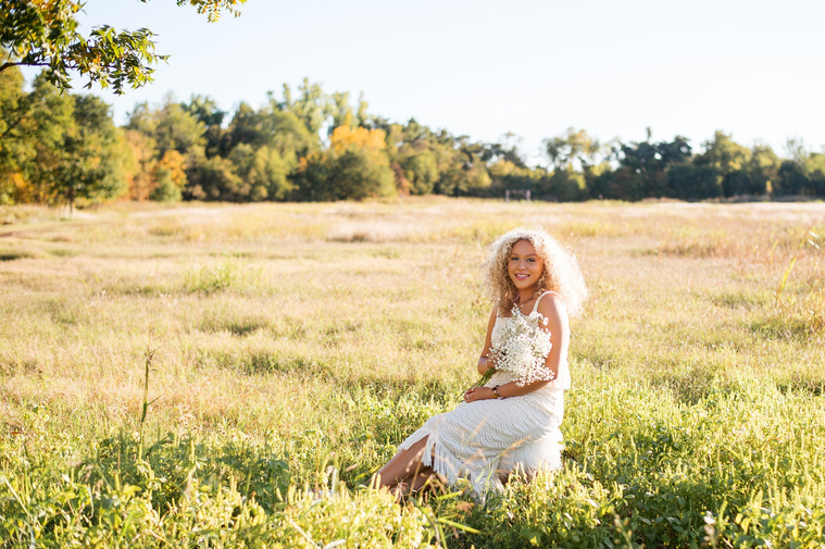wide angle portrait of a high school senior girl with blonde curly hair and blue eyes wearing a white fringe dress holding a bouquet of baby's breath and smiling while she sits on a stool in a field at Martin Nature Park in Oklahoma City