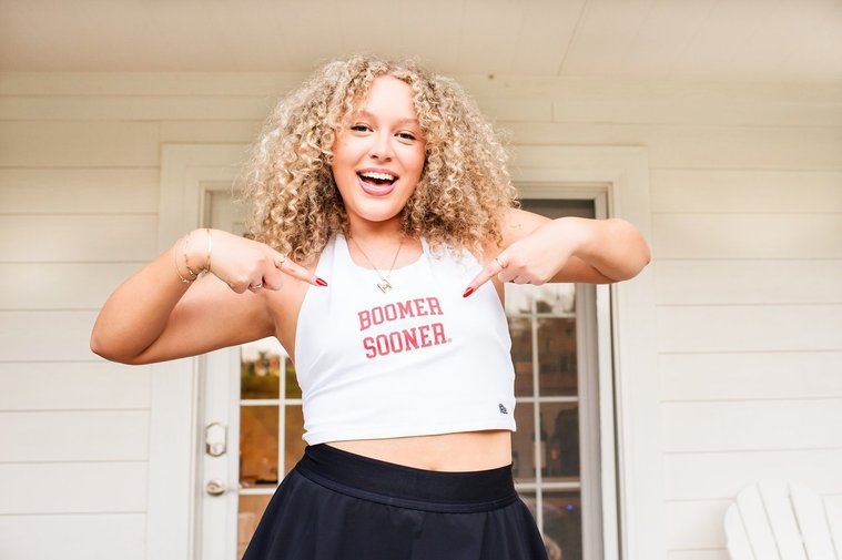 a senior girl wearing a white boomer sooner tank smiles and points at the words standing on her porch in Norman, Oklahoma
