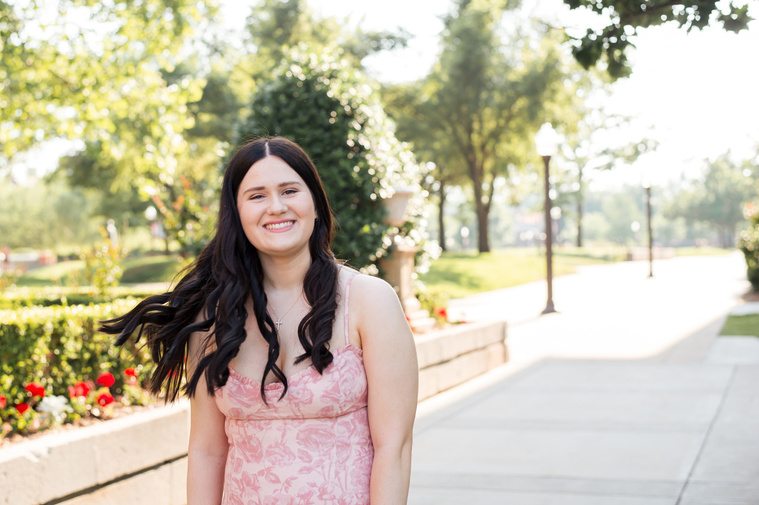 college senior girl wearing a light pink floral sundress smiles with her long dark hair flying around her while she stands on a sidewalk on OU campus in Norman
